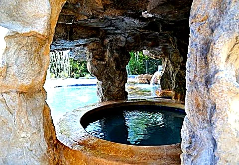 View from inside a carved concrete grotto over a round spa, looking out at the pool