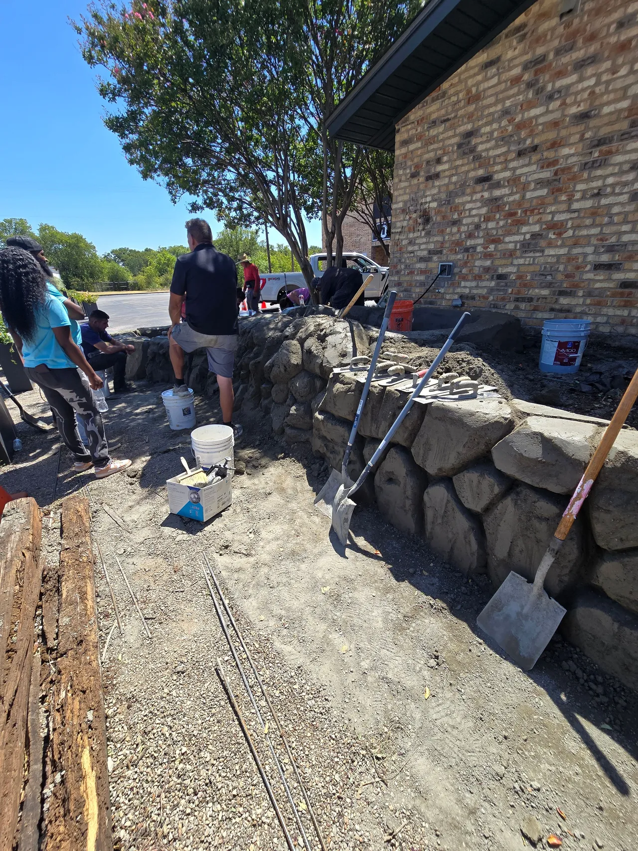 Old railroad ties pulled out and stacked beside a new hand-carved concrete boulder retaining wall going up in Burleson TX